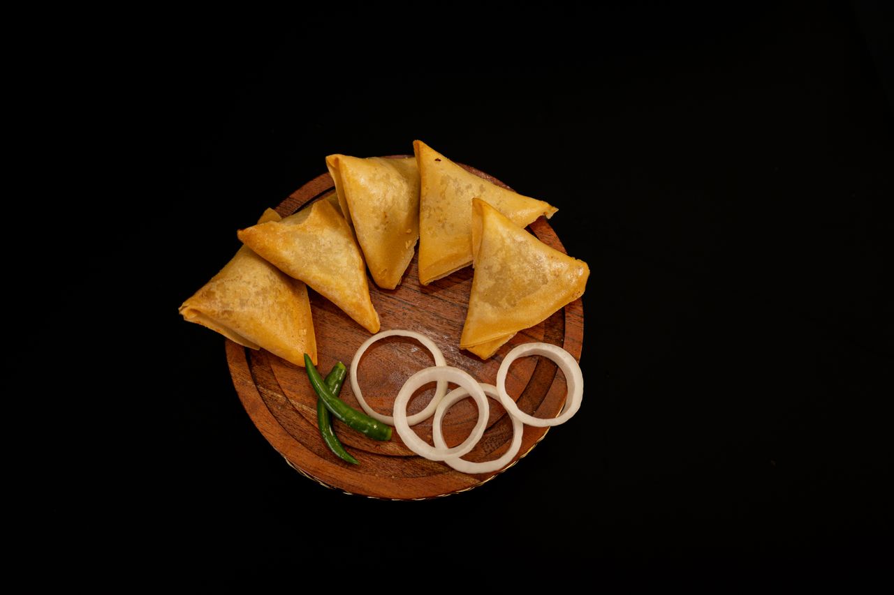 Four crispy samosas with onion rings and green chili on a wooden plate against black background.