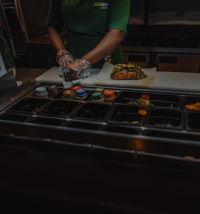 A Subway employee prepares a fresh sandwich with various ingredients at a fast-food restaurant.